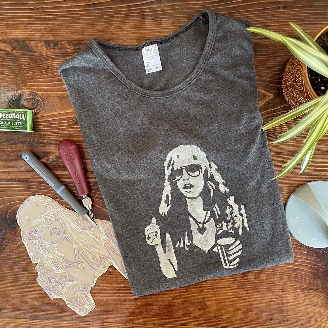A charcoal grey t-shirt with a white, hand-printed printed portrait of Stevie Nicks, folded on a wood background with linocut tools surrounding it.